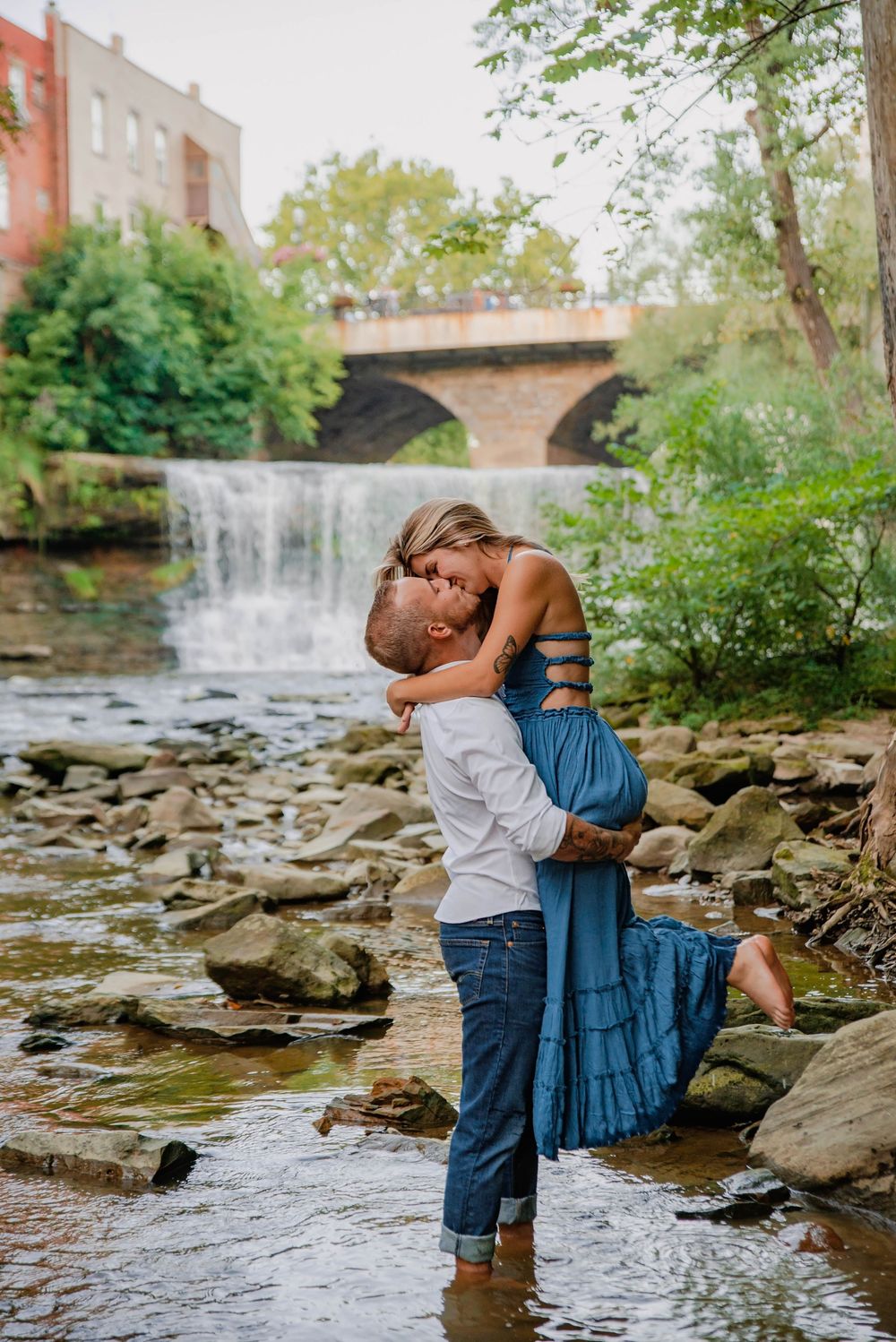 Romantic couple photos by a rocky stream with a waterfall backdrop showing playful interaction.
