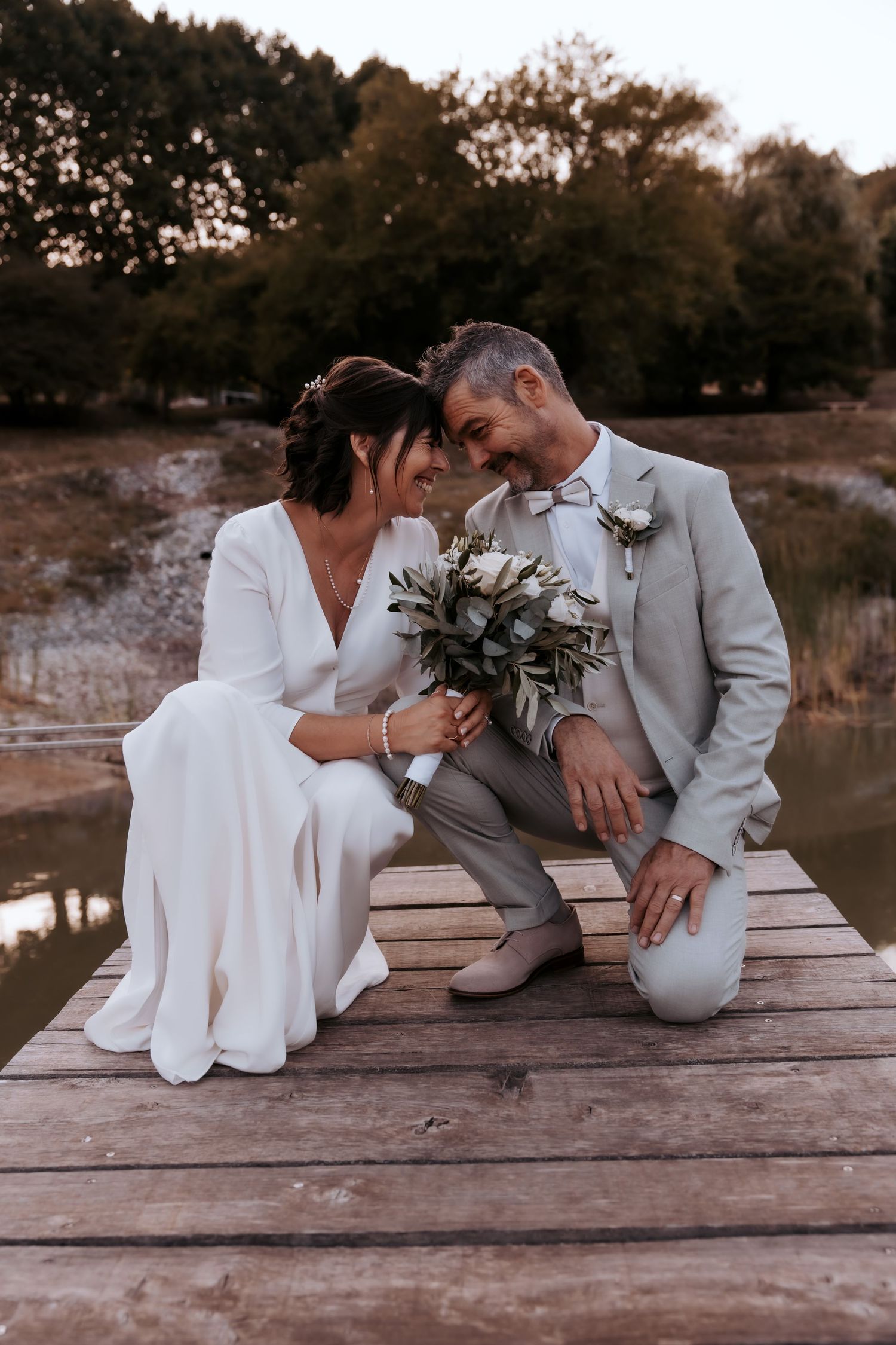 A couple in wedding attire sitting intimately on a wooden dock by a lake.Domaine la Plume photographe mariage