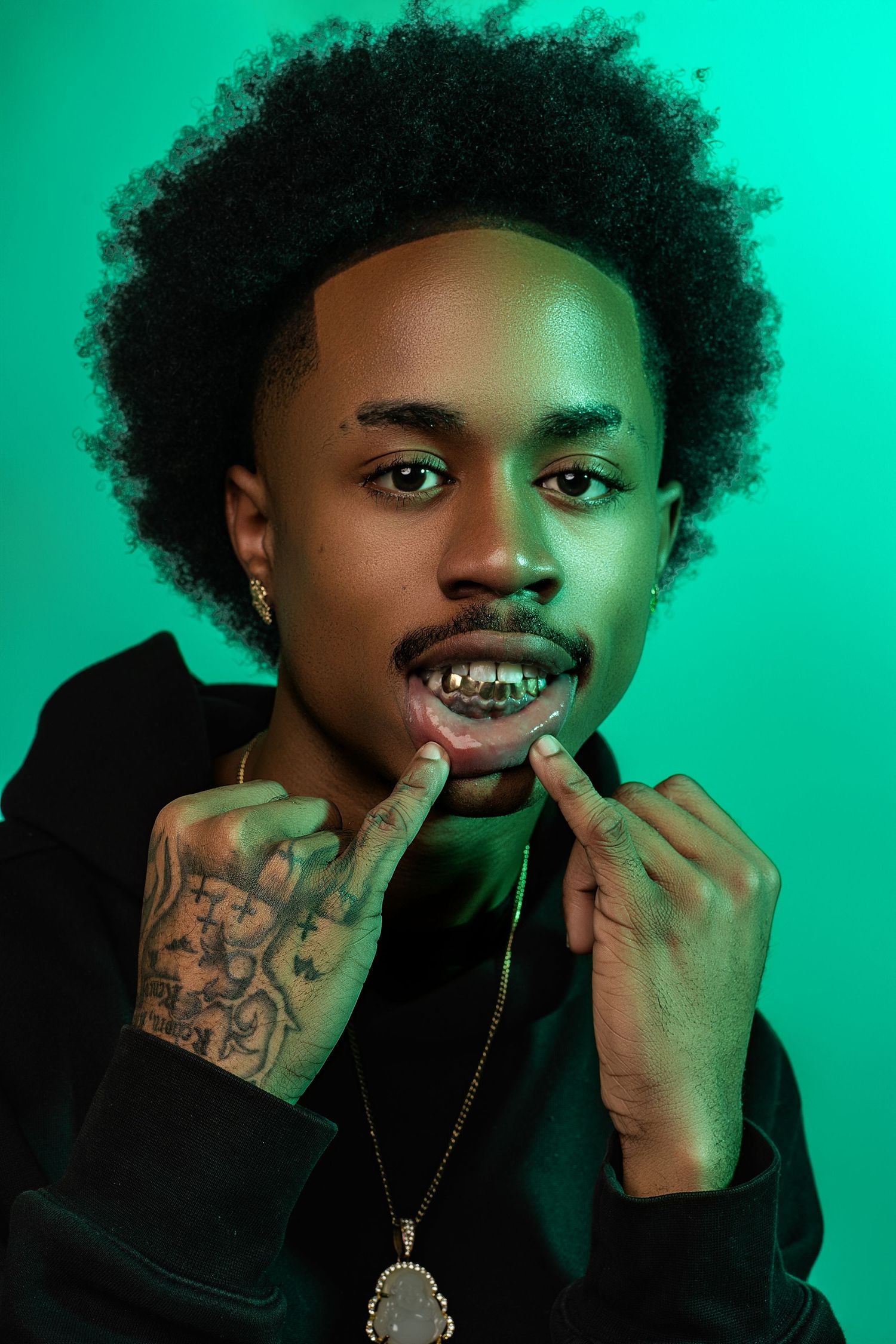 Person with afro hair crouches on stool wearing black t-shirt and pants against vibrant green backdrop in studio portrait.