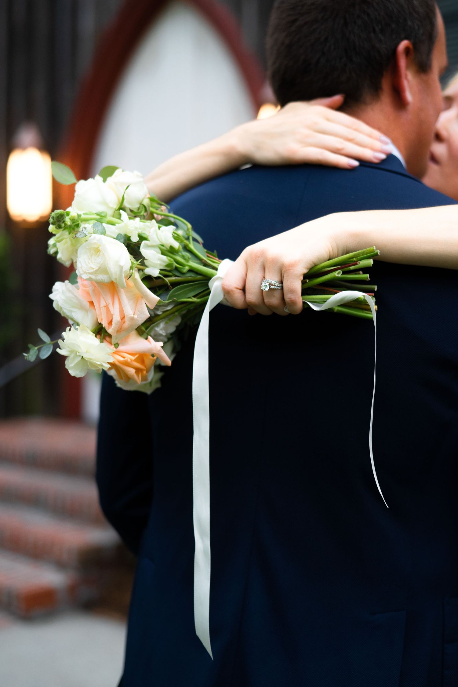A bouquet of peach and white roses with trailing white ribbons is held against a navy blue background.