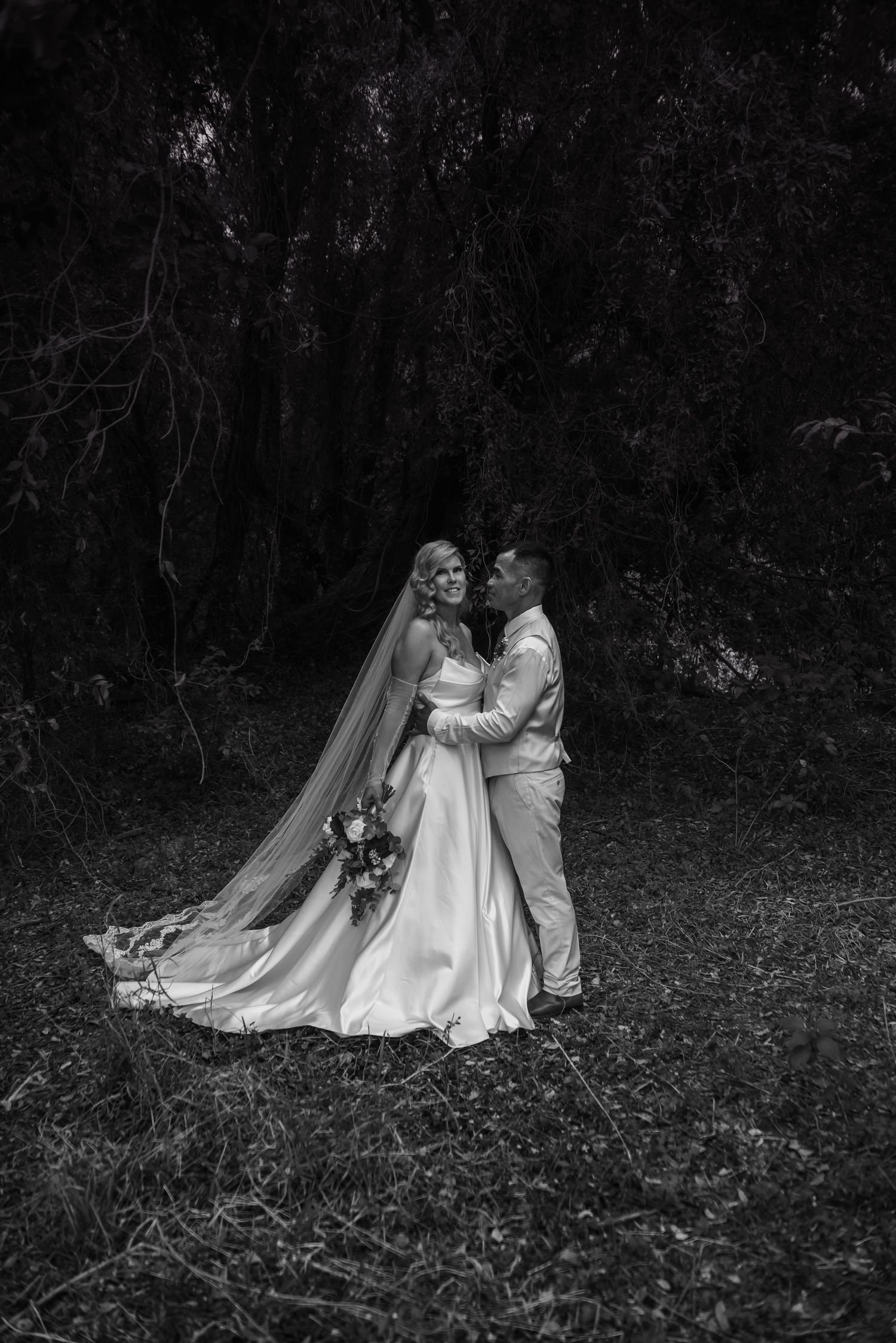 A black and white photograph shows a couple in wedding attire standing together in an open field at dusk.