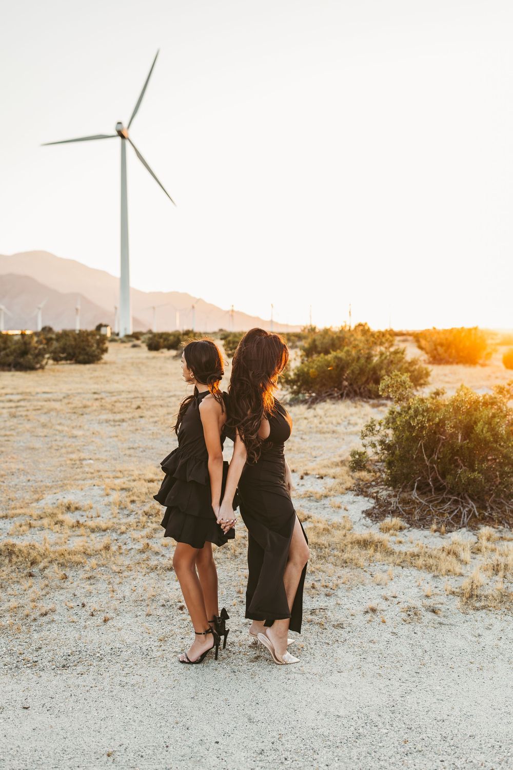 Two people in black dresses share a tender moment at sunset with wind turbines in the desert landscape.
