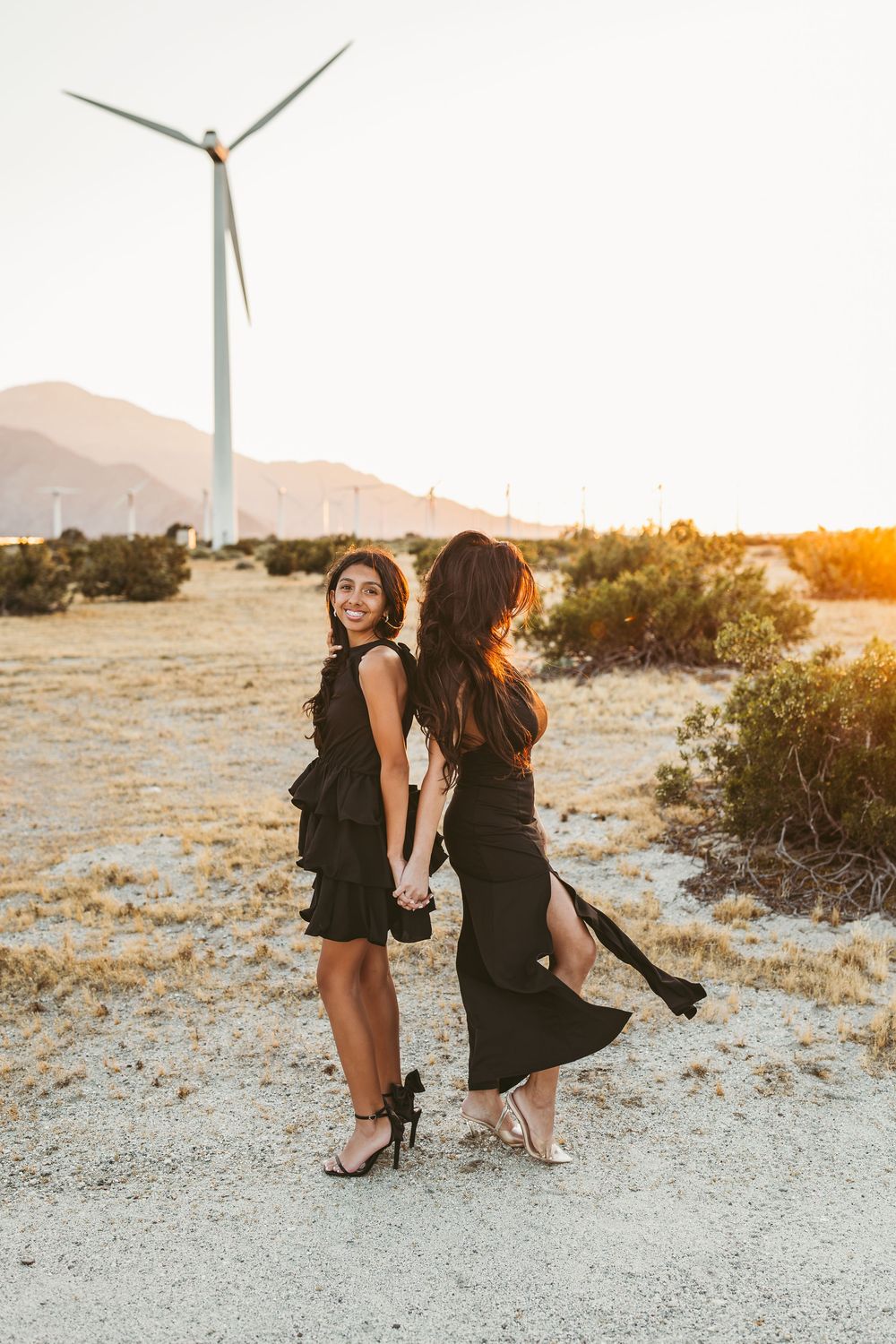 Two friends in black outfits laugh together in a desert setting with wind turbines at golden hour.