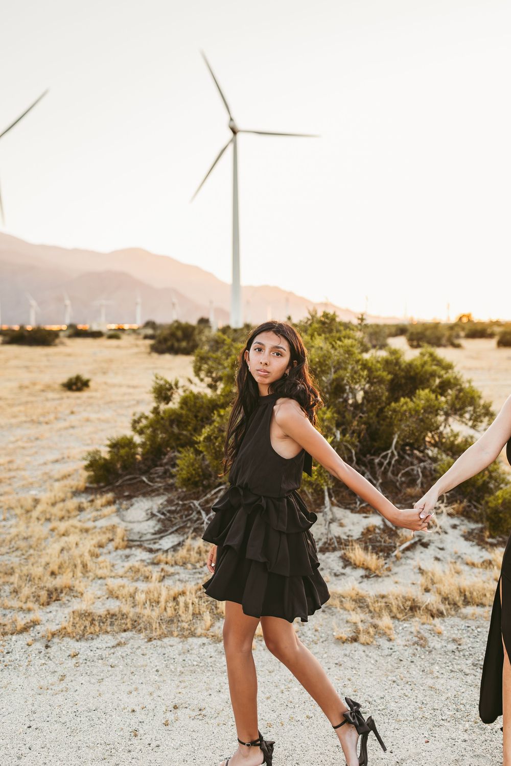 A figure in a black dress poses gracefully against a desert landscape with wind turbines in the background at sunset.