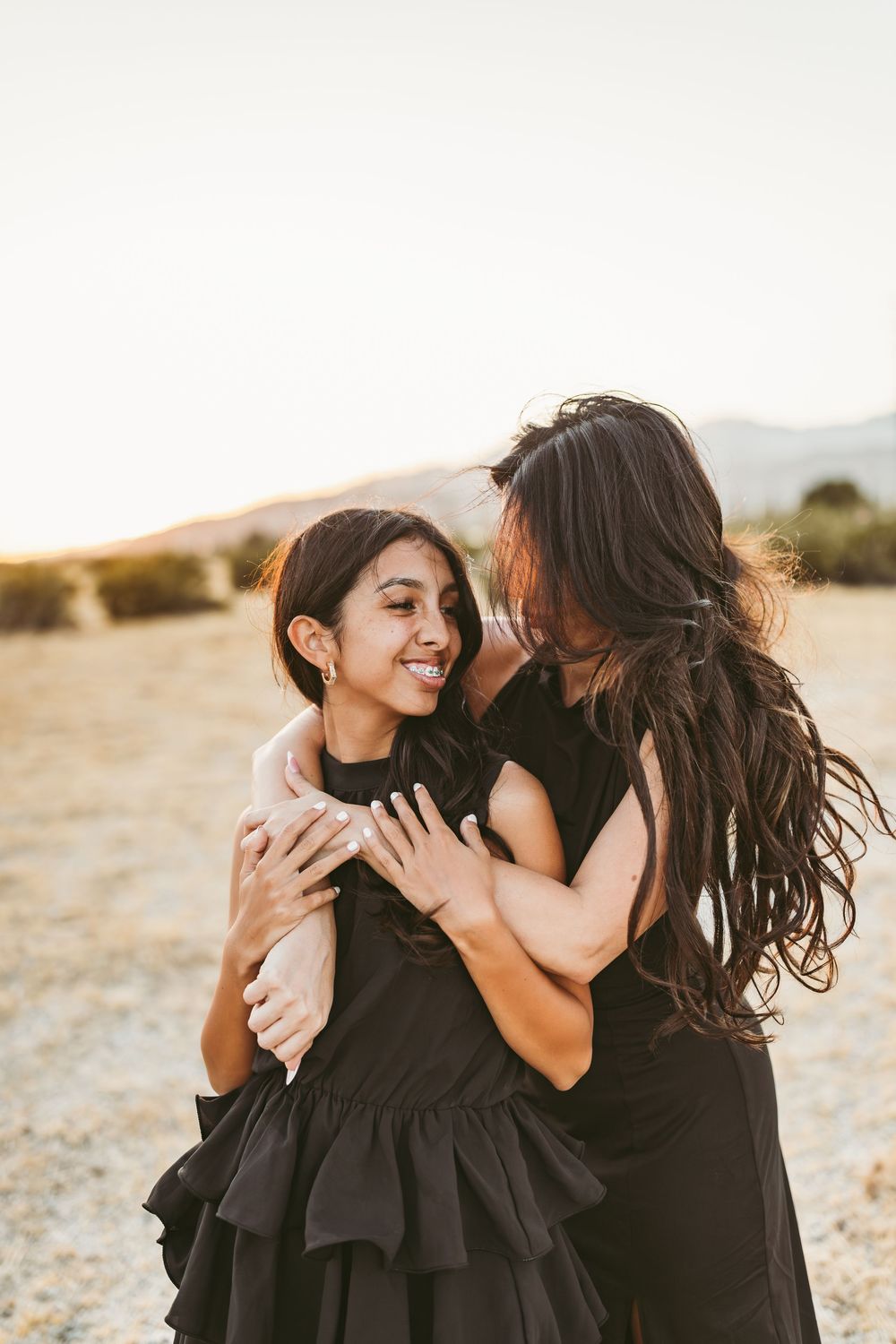 Two people in black dresses embrace joyfully against a warm sunset backdrop in the desert.