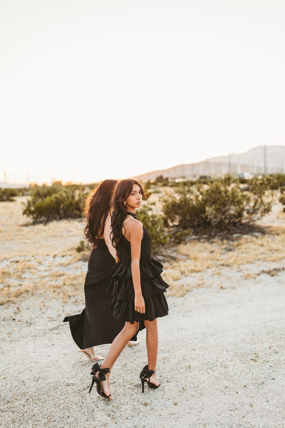 Two people in black dresses pose playfully on desert terrain at sunset with mountains in the background.