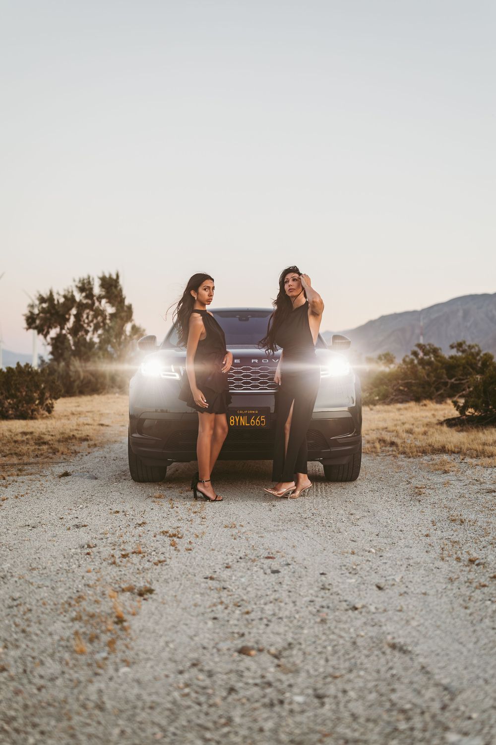 Two women in black dresses pose in front of a luxury SUV with headlights on at sunset in a desert setting.