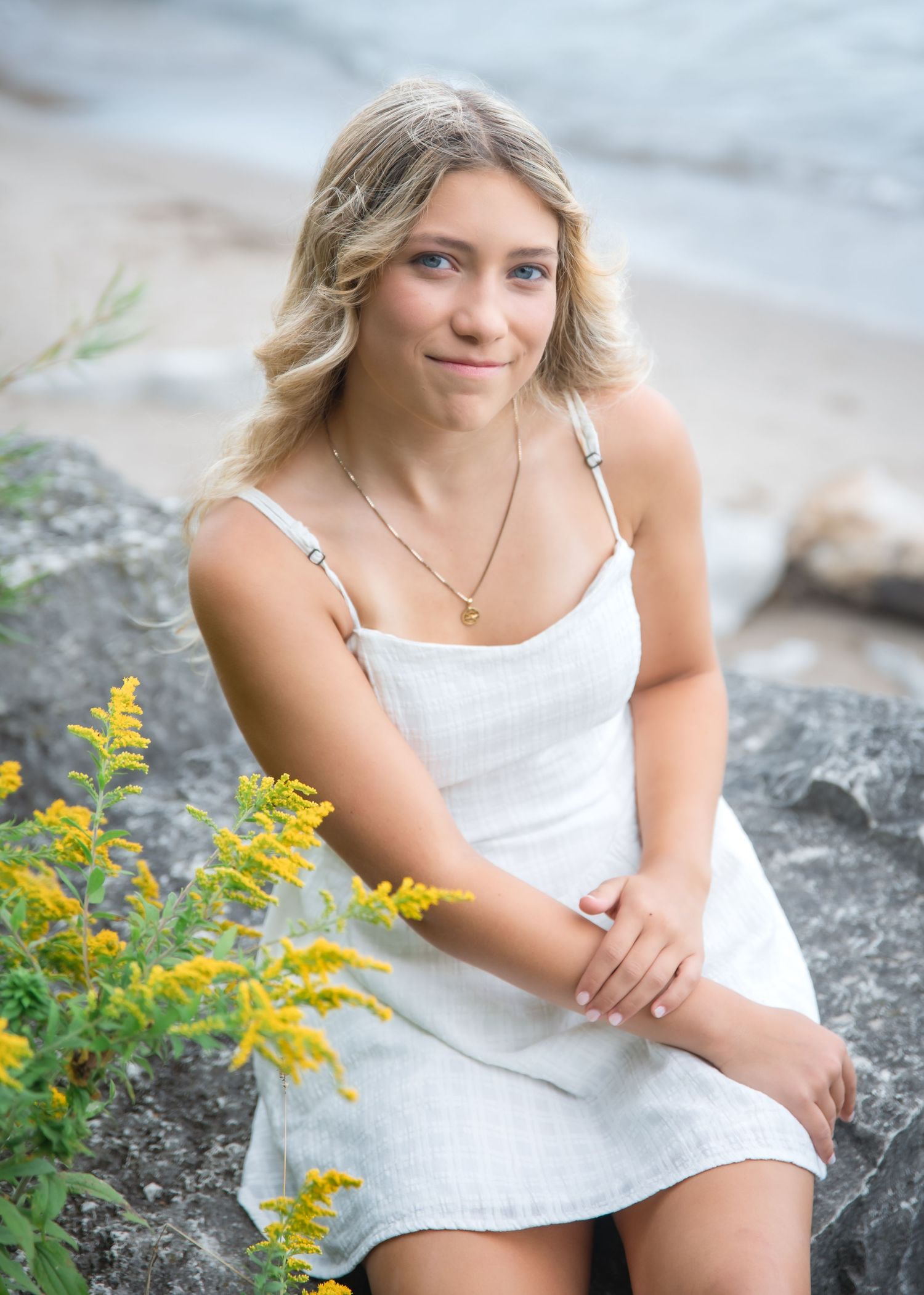 Young blonde woman in white sundress sitting on rocks near beach with yellow wildflowers in foreground.