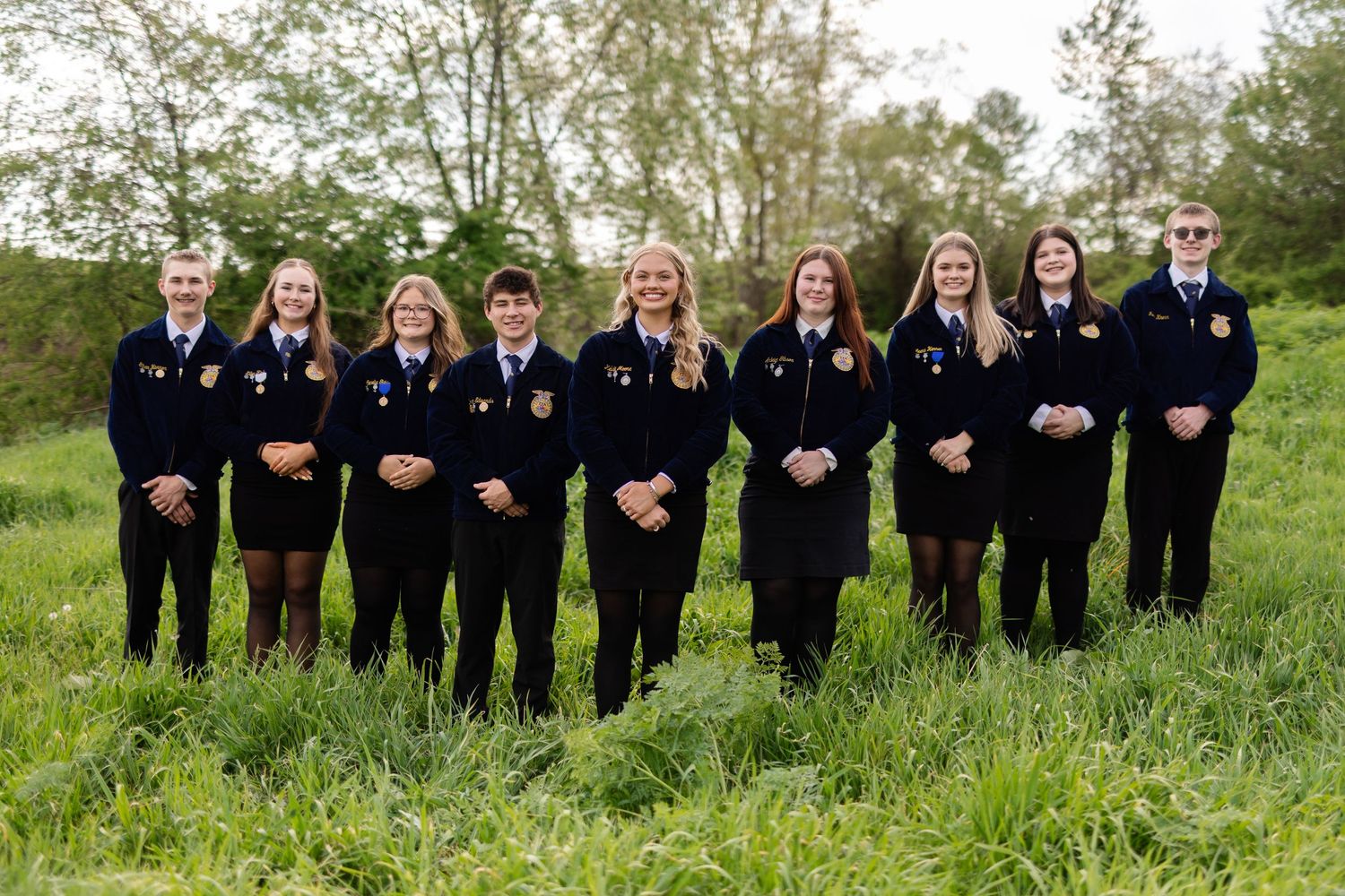 Nine students in matching FFA black jackets pose together smiling outdoors on a green grassy field.