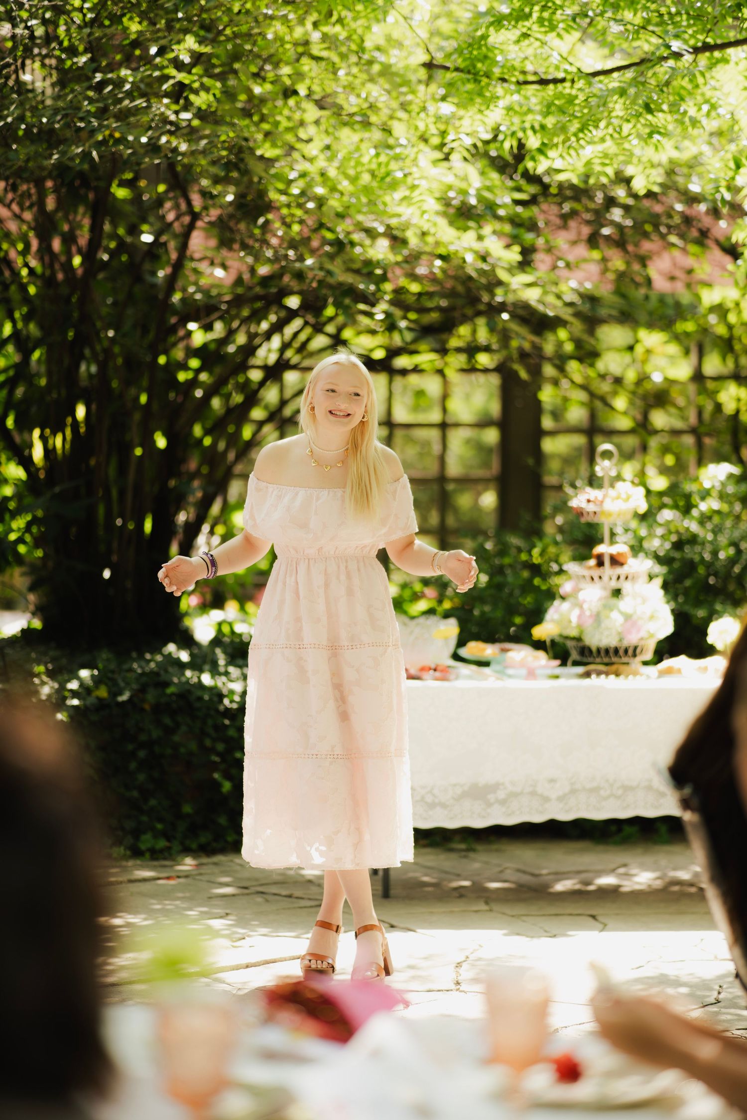 Smiling blonde woman in pink off-shoulder midi dress at outdoor garden party with tiered cake stand.