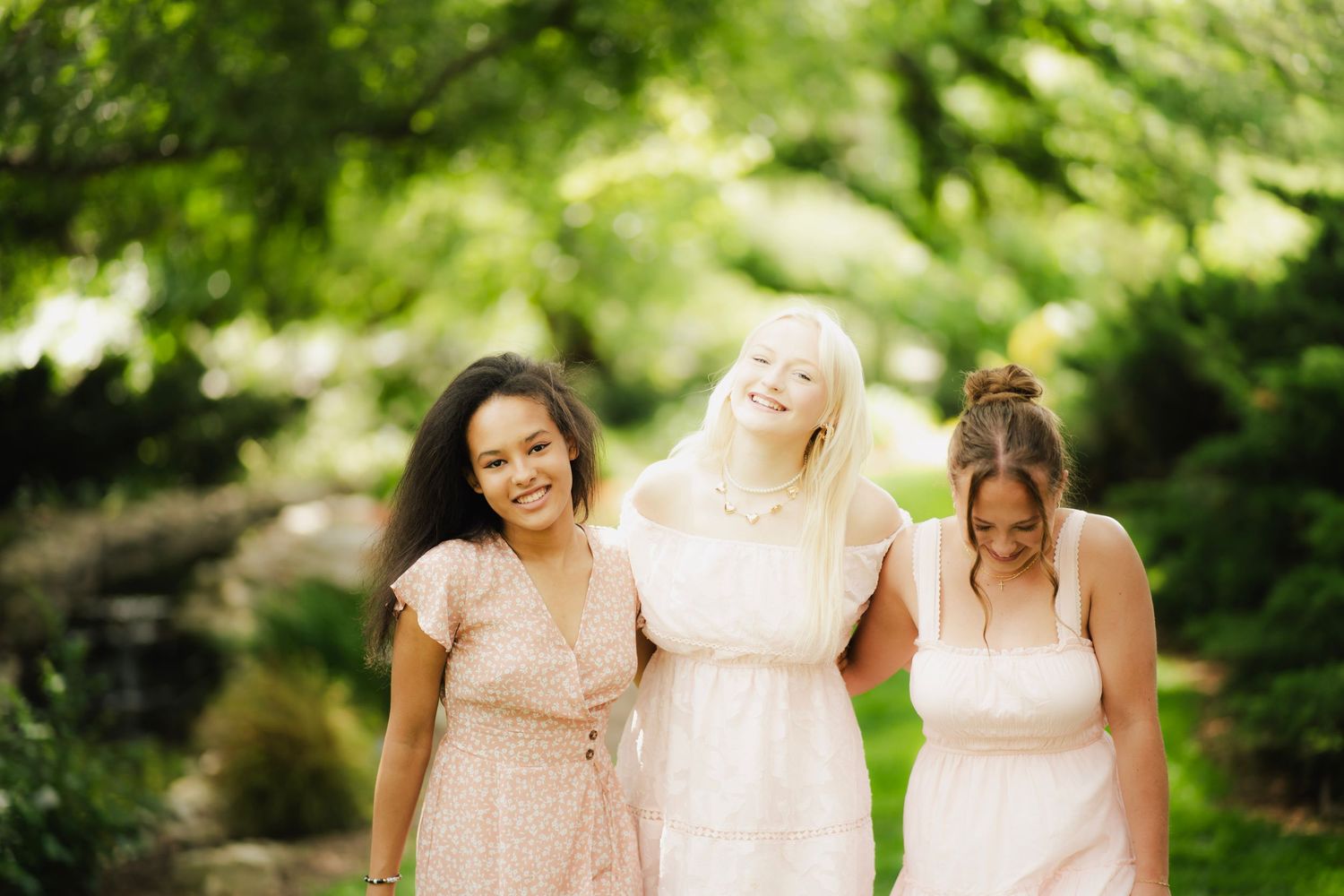Three women in pink and blush dresses smiling and walking together in a lush green garden.