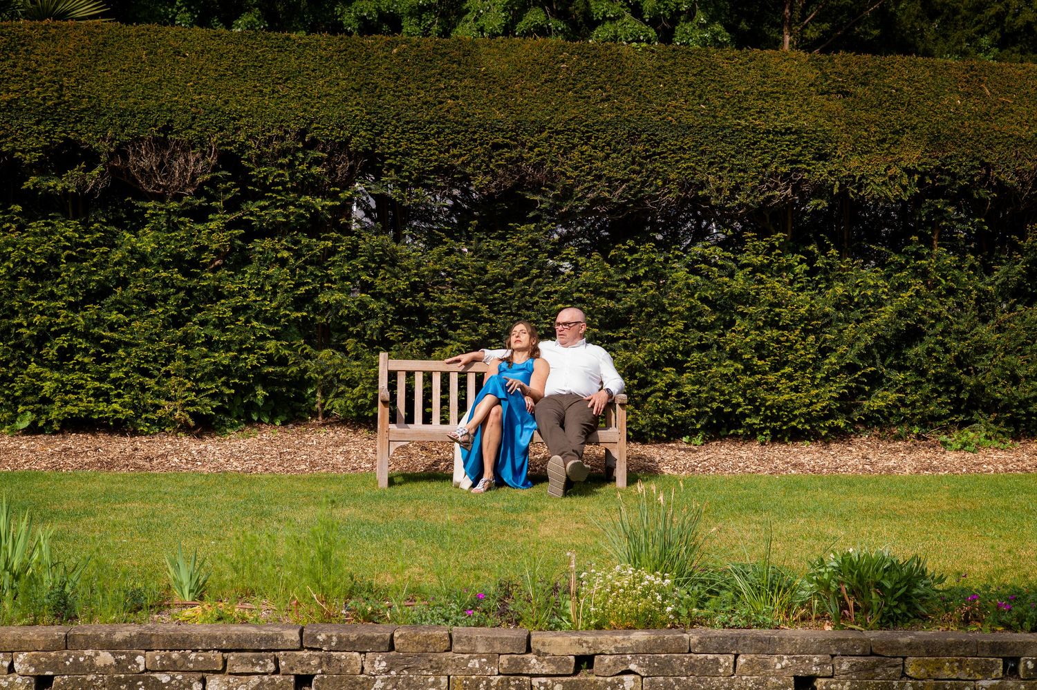 Couple sitting together on a wooden bench in a lush green garden with a tall hedge backdrop.
