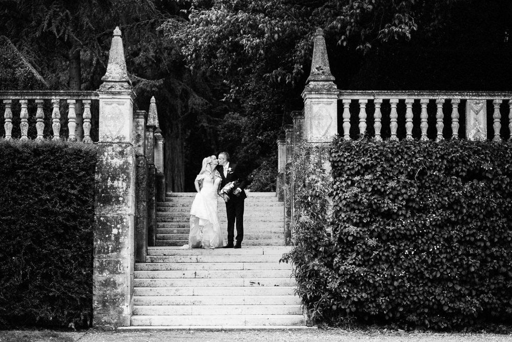 Black and white photo of a bride and groom walking hand in hand down a lush garden pathway on their wedding day.