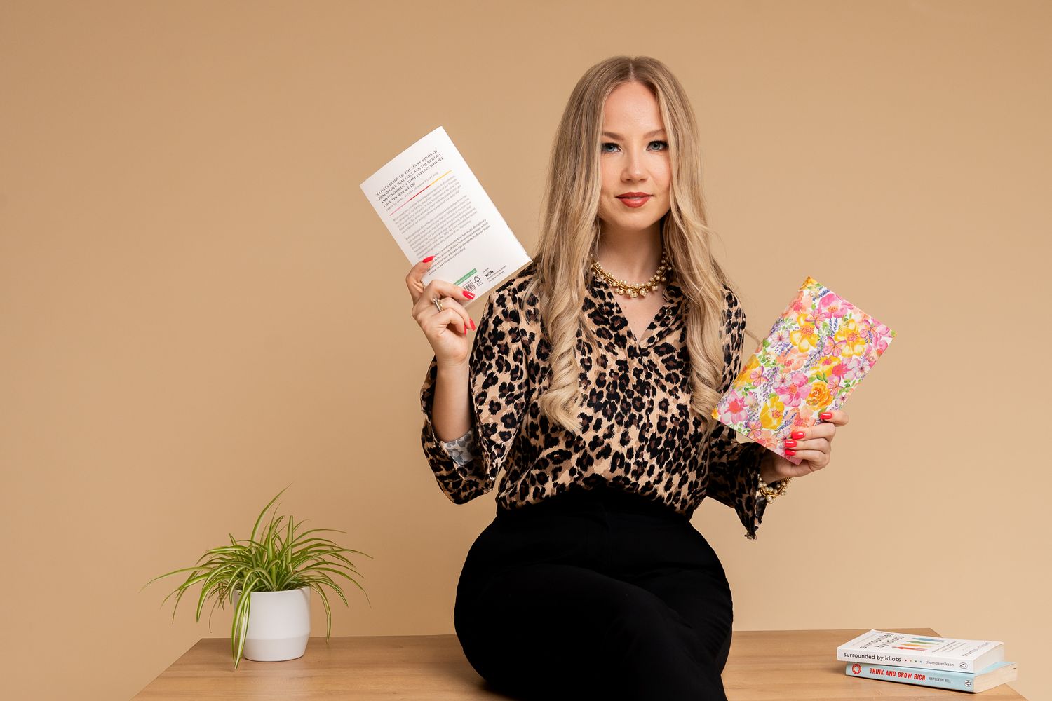 A business woman in a leopard print blouse sitting on desk holding documents and colorful notebook against beige background. Cambridge, UK