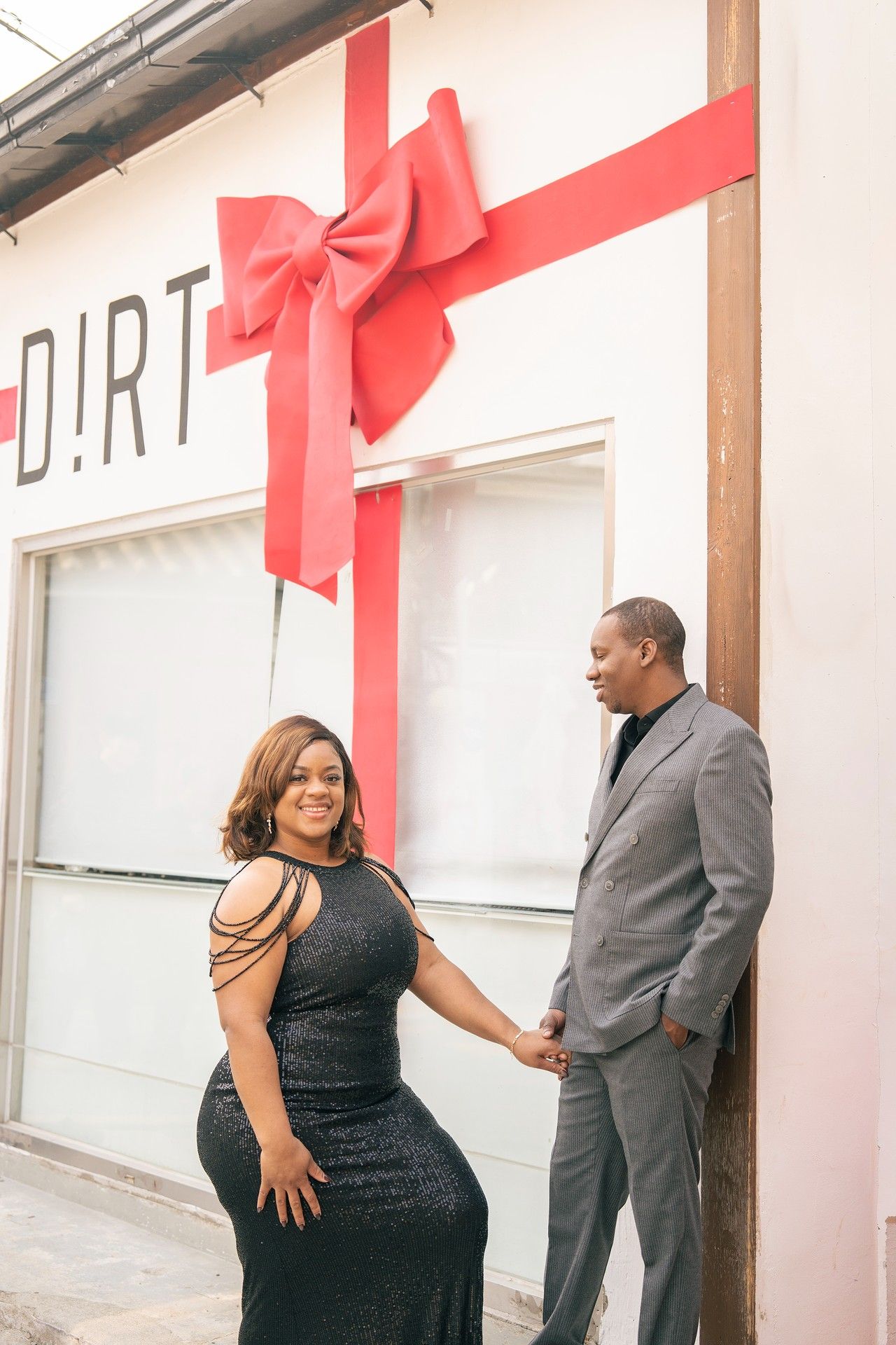 Couple in elegant attire holding hands in front of a white building decorated with a large red bow.