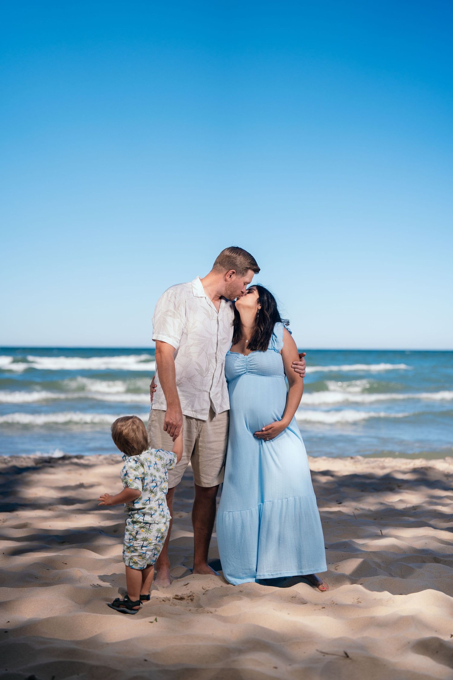 Pregnant woman in blue dress with husband and toddler on sunny beach, family maternity photo session by the ocean.