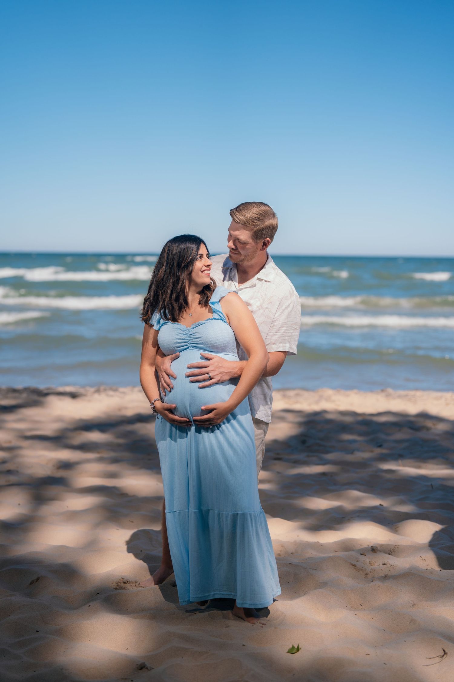 Pregnant couple embracing on a sunny beach, woman in blue maxi dress, man in white shirt, ocean waves behind them.