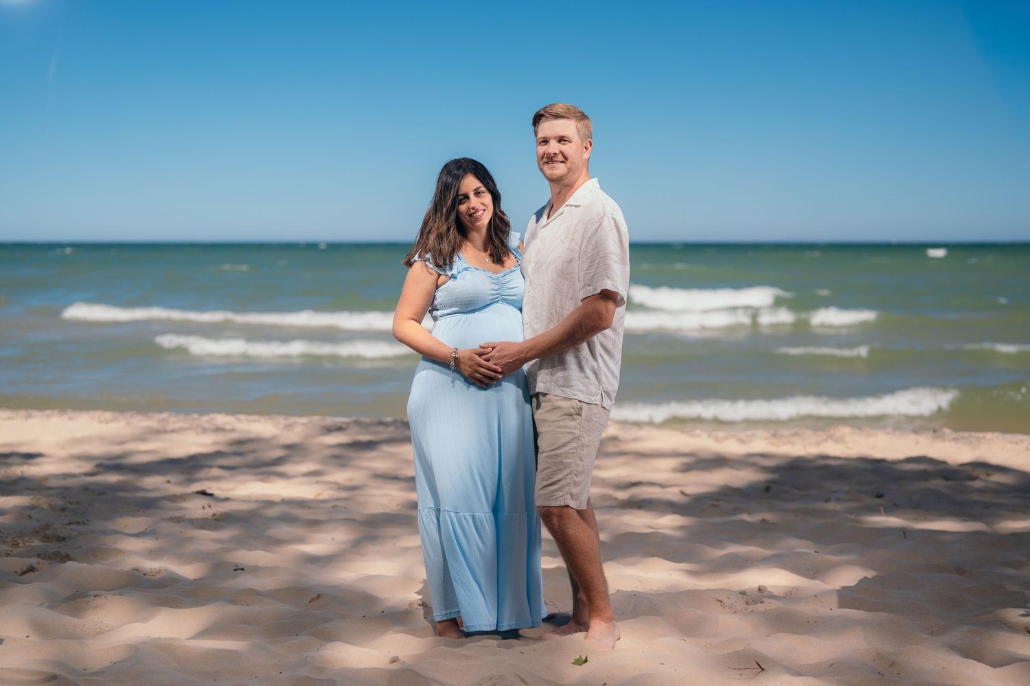 Pregnant couple posing on sandy beach with ocean waves, woman in blue maternity dress, man in casual summer outfit.
