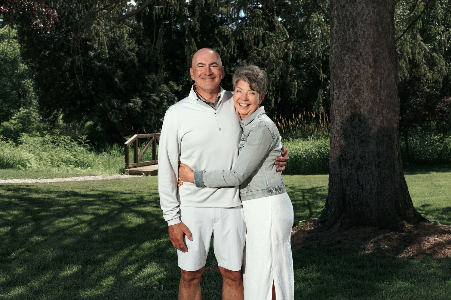 Smiling older couple embracing outdoors on a green lawn near a large tree on a sunny day.