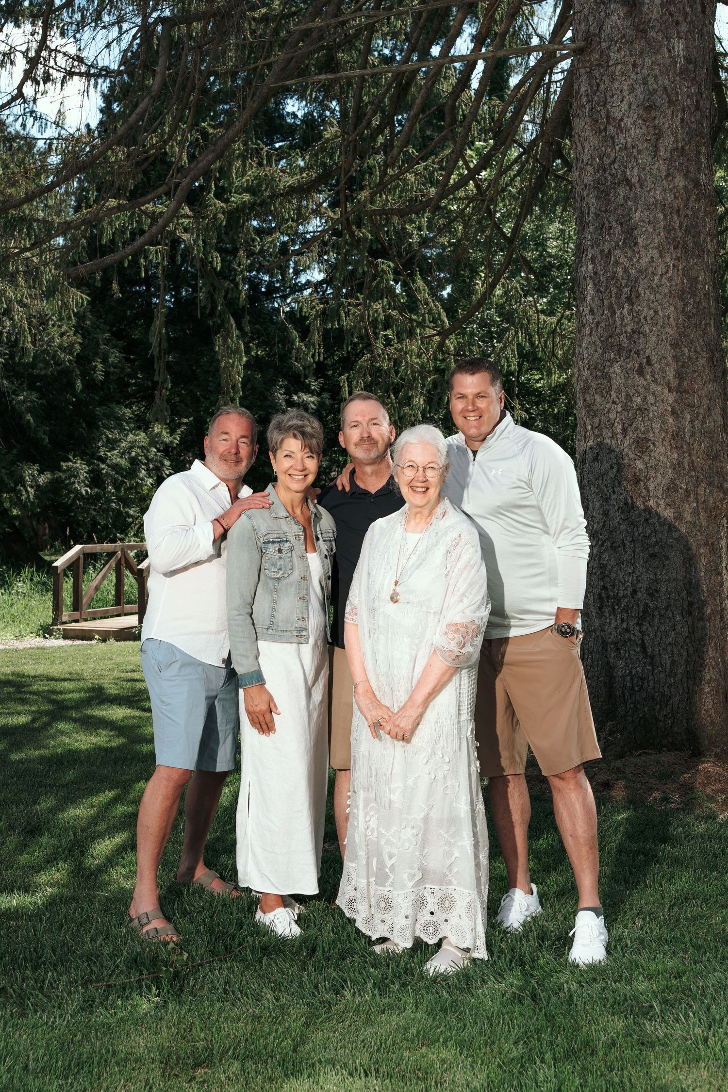 Family of five smiling outdoors under a large tree on a sunny day in a lush green park setting.