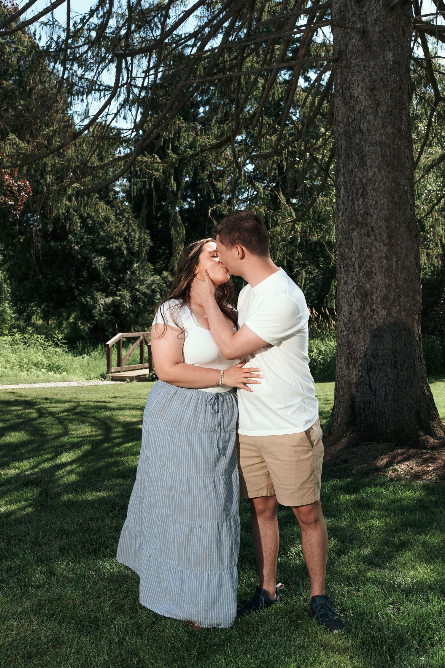A couple shares a romantic kiss under a large tree in a lush green park, woman in gray maxi dress.