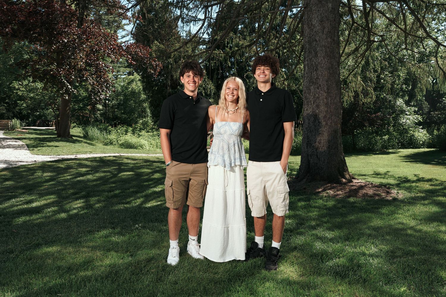 Three young people posing outdoors in a lush green park, woman in white dress flanked by two men in casual attire.
