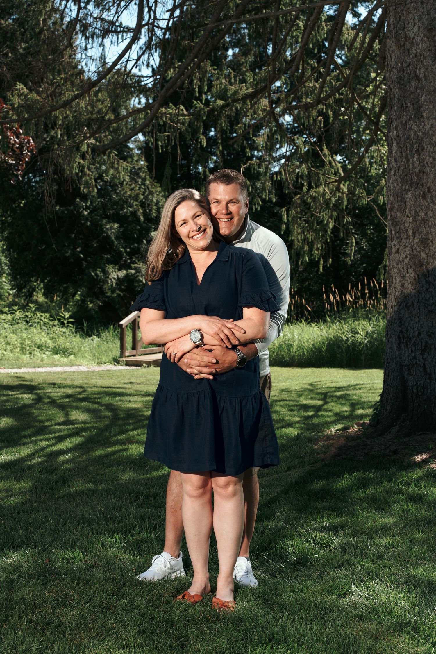 Happy couple embracing outdoors in a lush green park, woman in navy dress, man hugging her from behind, smiling.
