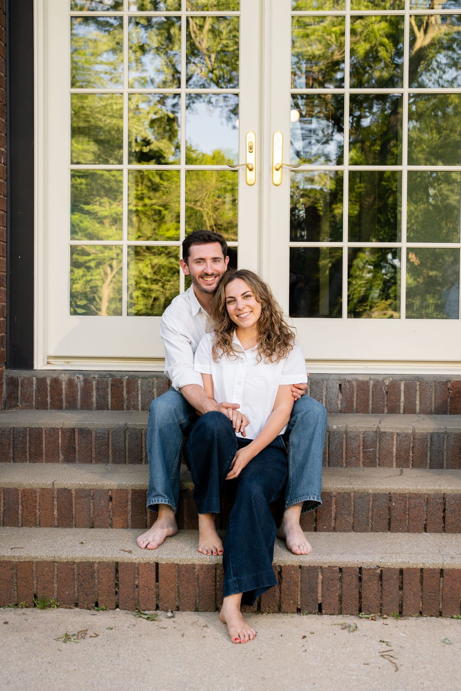 Couple sitting barefoot on brick steps in front of French doors, smiling warmly in casual white shirts and jeans.