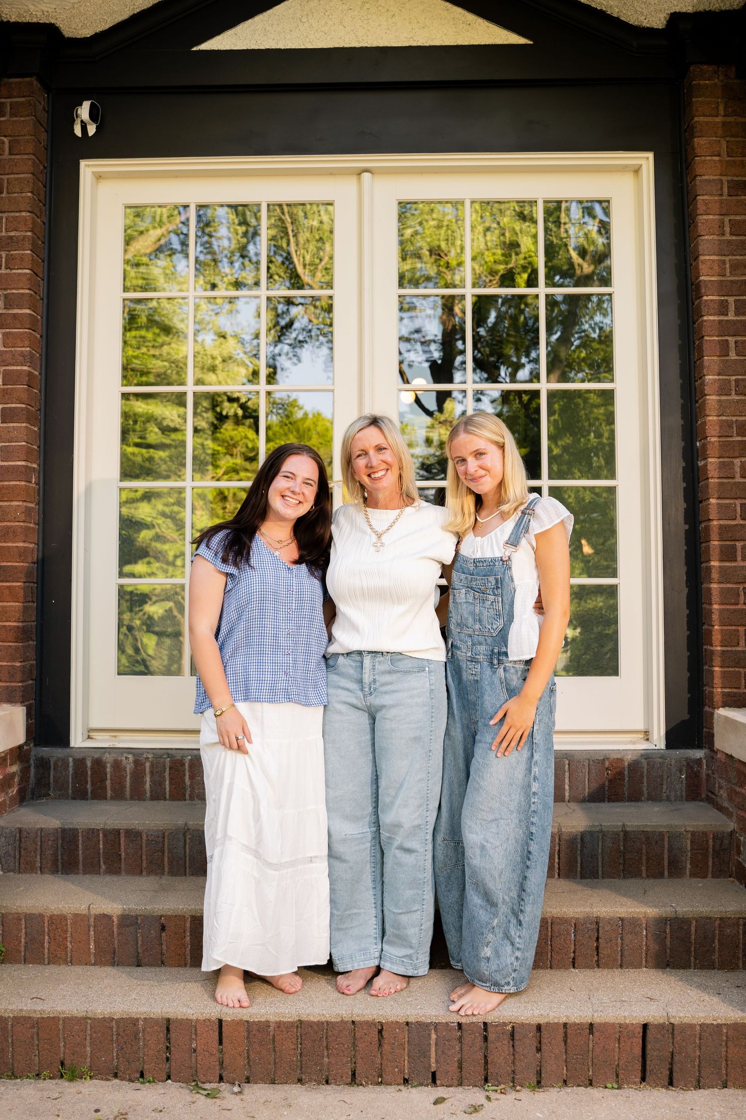 Three smiling women pose together on brick steps in front of a home with large windows on a sunny day.