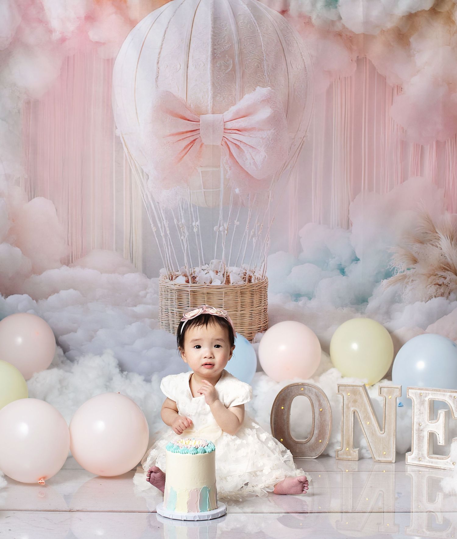 Baby girl in white dress sits by a smash cake, surrounded by pastel balloons and a hot air balloon backdrop.