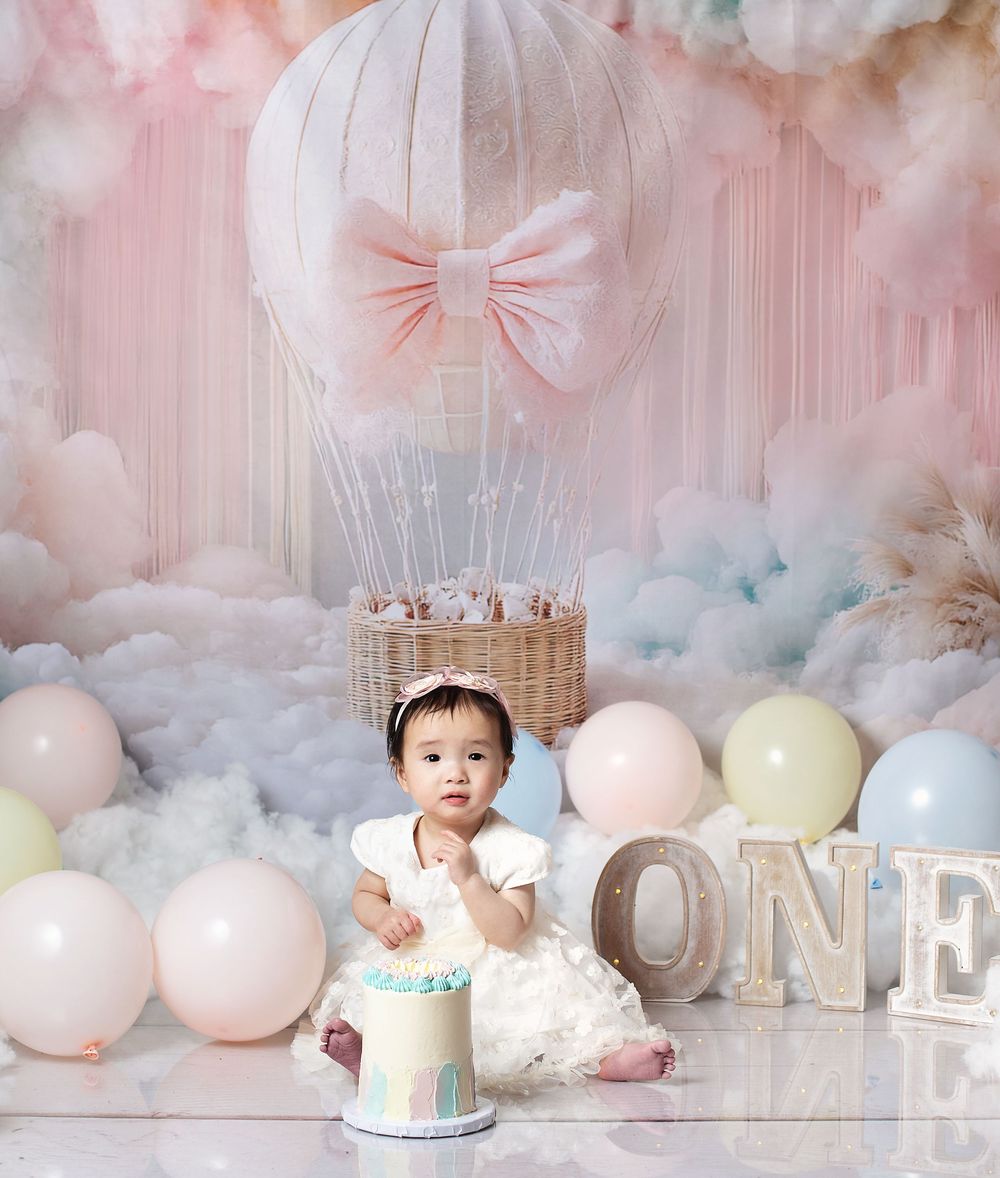 Baby girl in white dress sits by a smash cake, surrounded by pastel balloons and a hot air balloon backdrop.