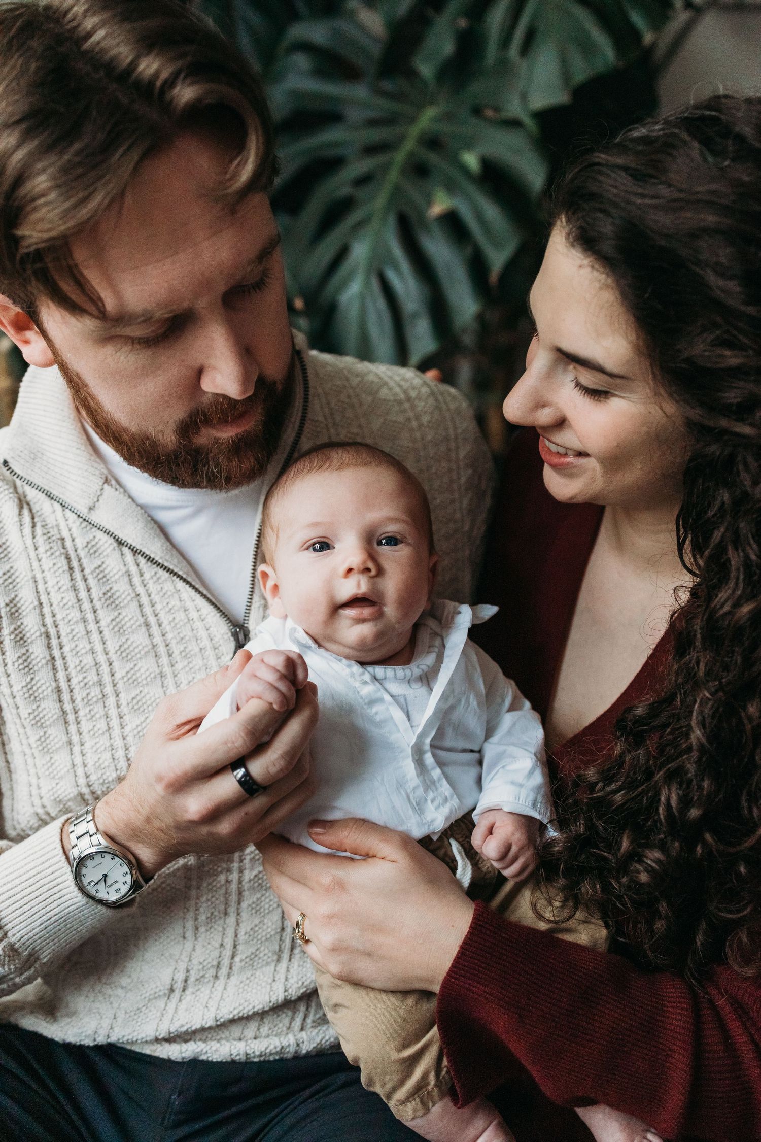 A bearded father and curly-haired mother lovingly hold their newborn baby dressed in white.