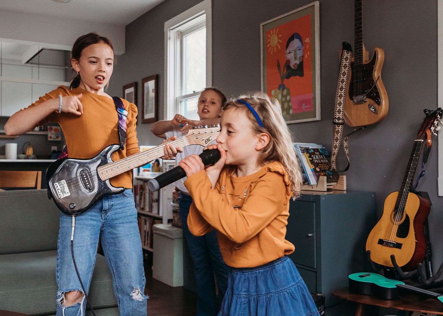Three girls playing music at home, two with guitars and one singing into a microphone, with guitars displayed on the wall.