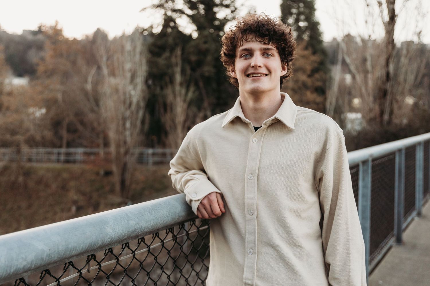 Smiling teenage boy with curly hair leaning on a bridge railing outdoors in casual beige shirt.