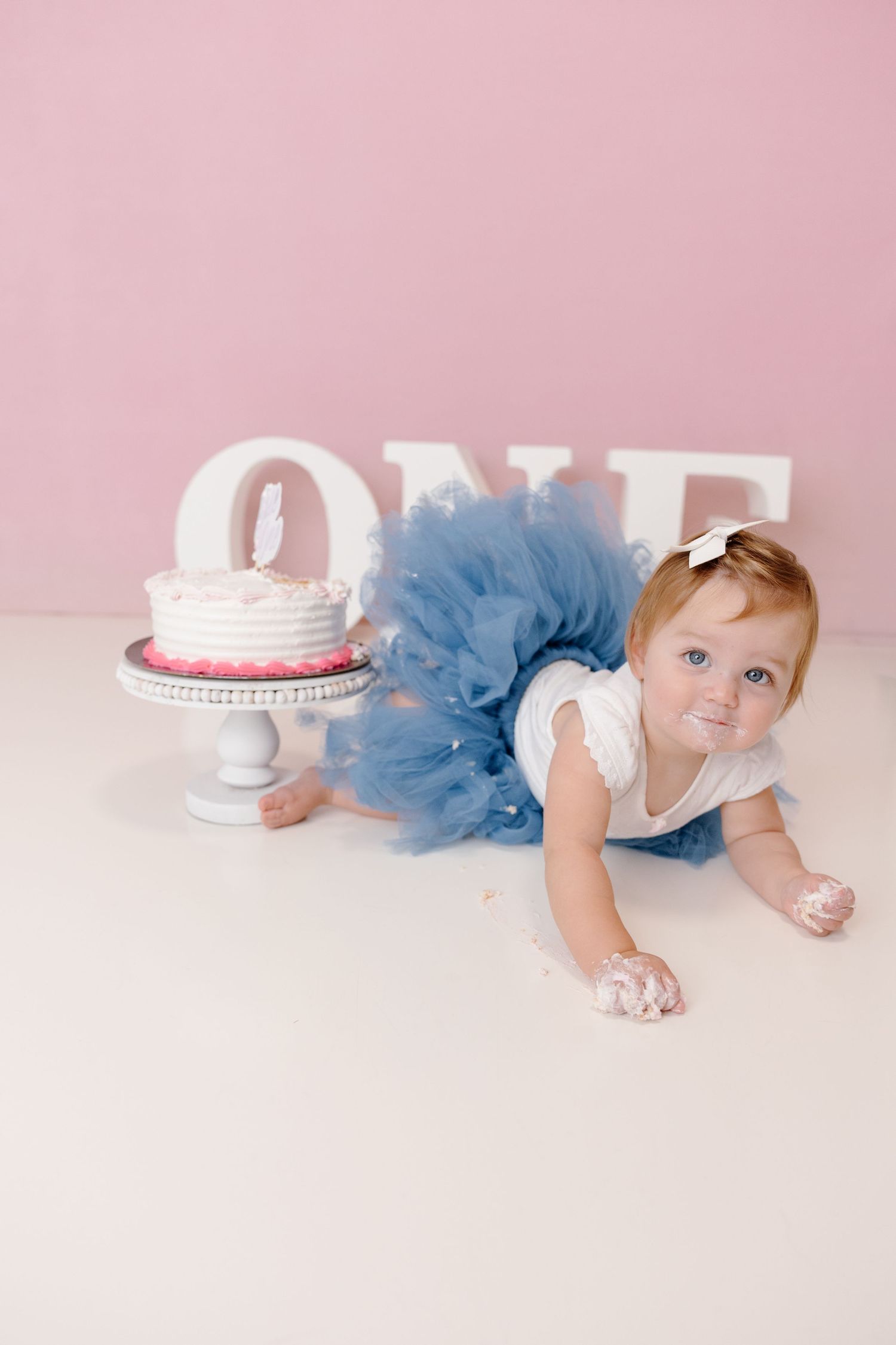 Baby girl in blue tutu crawls near smash cake during first birthday photo session with pink backdrop.