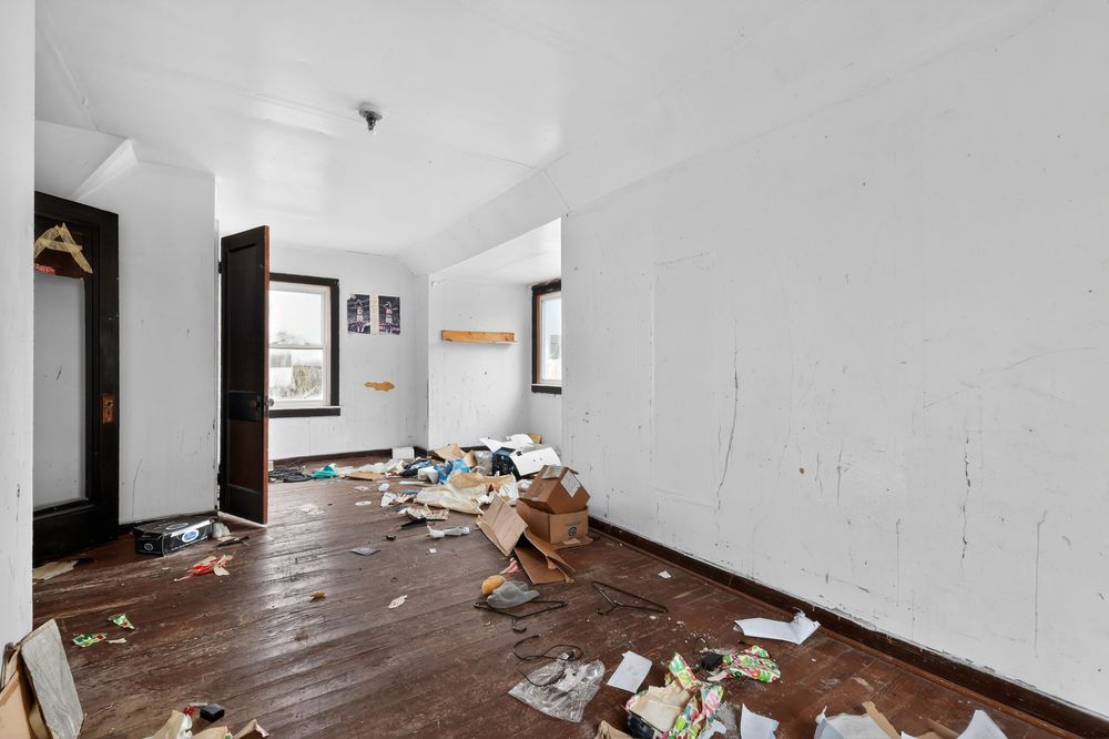 Abandoned room with scattered debris on worn hardwood floors, white walls, and natural light from windows. Detroit MI