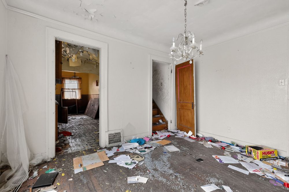 Abandoned room with debris-covered floor, peeling walls, crystal chandelier, and open doors revealing staircase. Detroit MI