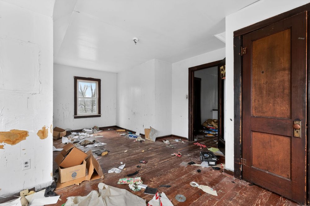 Abandoned room with debris-covered hardwood floors, white walls, a wooden door, and a window letting in natural light.