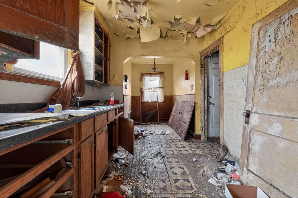 Abandoned kitchen with peeling ceiling, damaged cabinets, debris-covered floor, and deteriorating walls in a derelict house.
