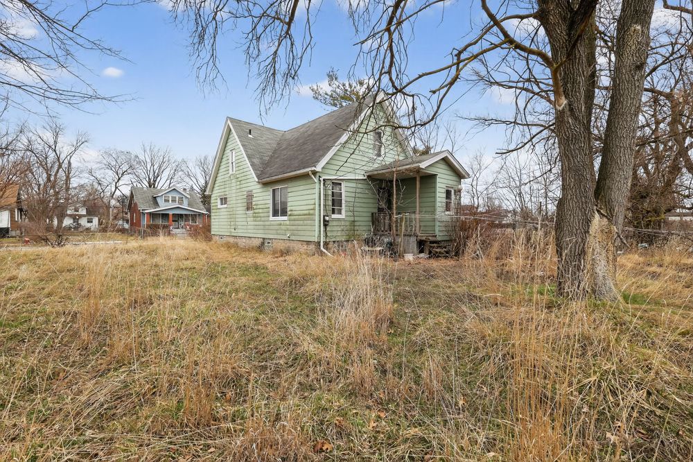 A weathered green bungalow with a front porch sits amid overgrown grass and bare trees under a cloudy sky. Detroit MI