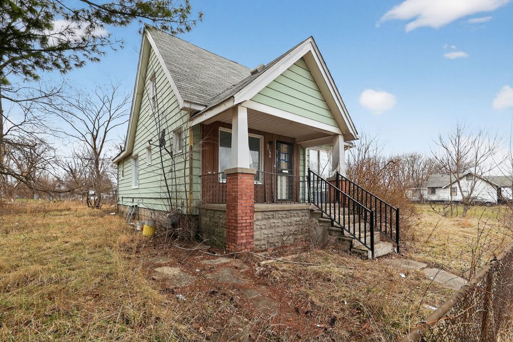 Abandoned green bungalow with brick porch columns, overgrown yard, and bare trees on a cloudy day. Detroit MI