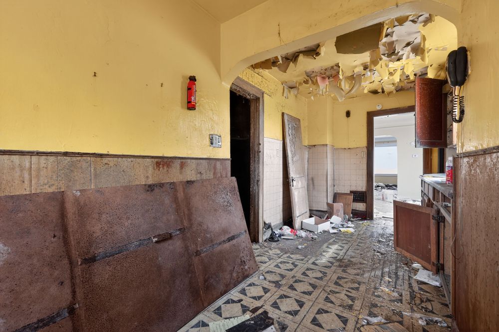 Abandoned building interior with damaged walls, peeling paint, debris on tiled floor, and a red fire extinguisher. Detroit MI
