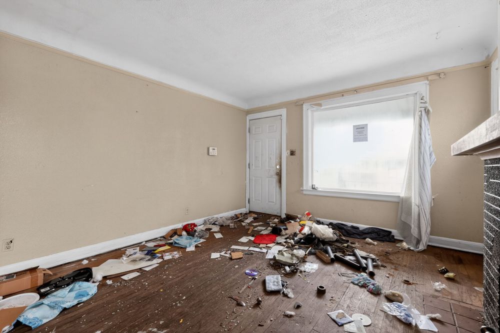 Abandoned room with debris and trash scattered across worn hardwood floors, damaged walls, and a window with torn curtains. Detroit MI