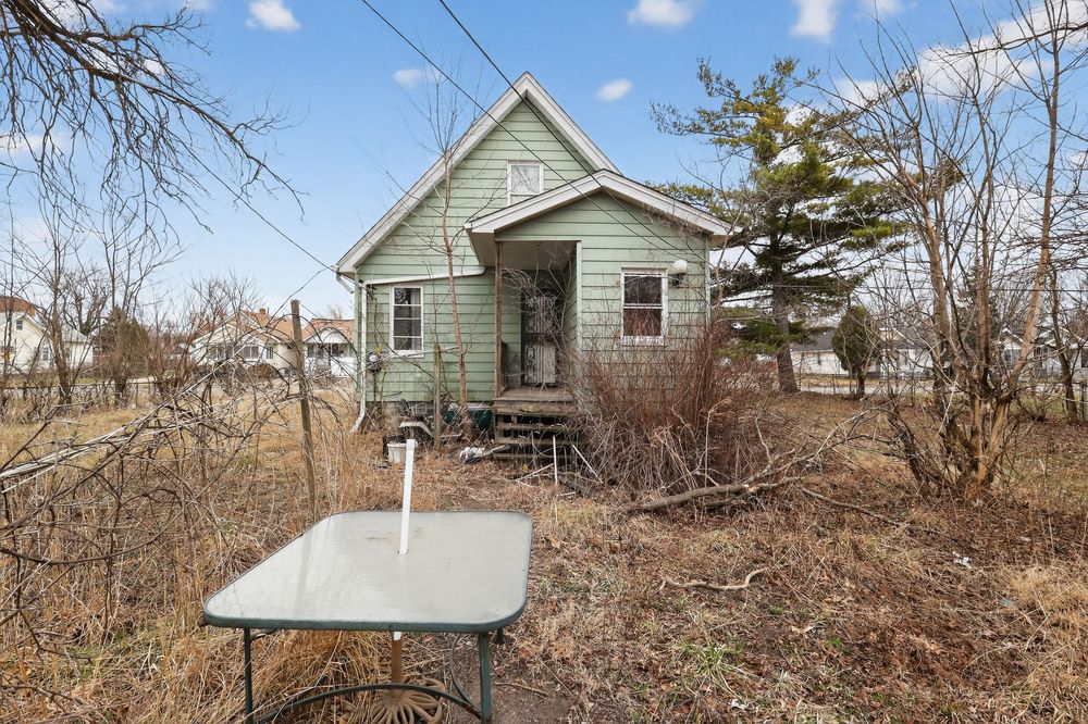 Small green vintage cottage with overgrown yard, wooden porch steps, and bare trees in early spring. Detroit MI