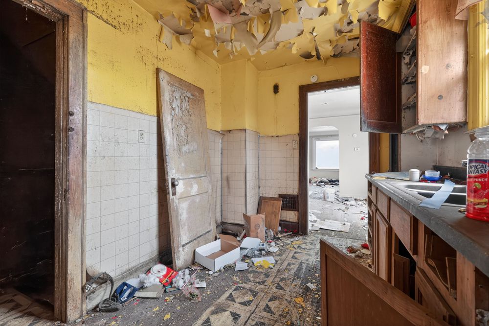 Abandoned kitchen with peeling yellow walls, debris-covered floor, damaged cabinets, and deteriorating ceiling in derelict building. Detroit