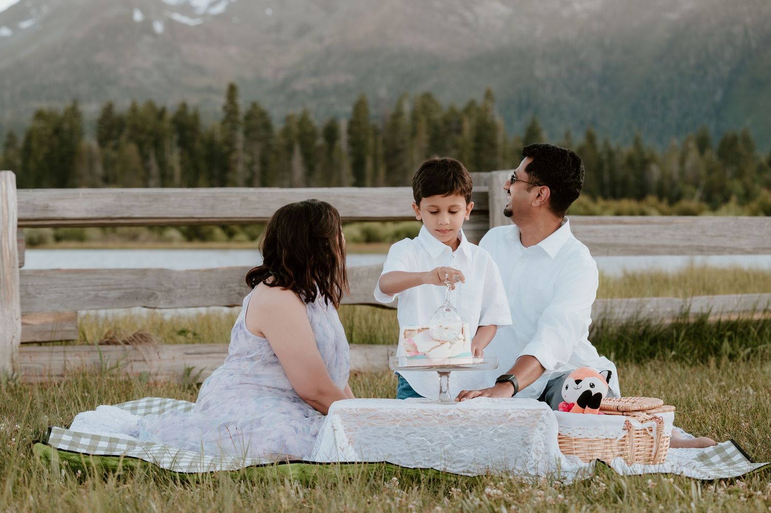 Family of three enjoying a outdoor picnic in a mountain meadow, with a young child cutting a cake.