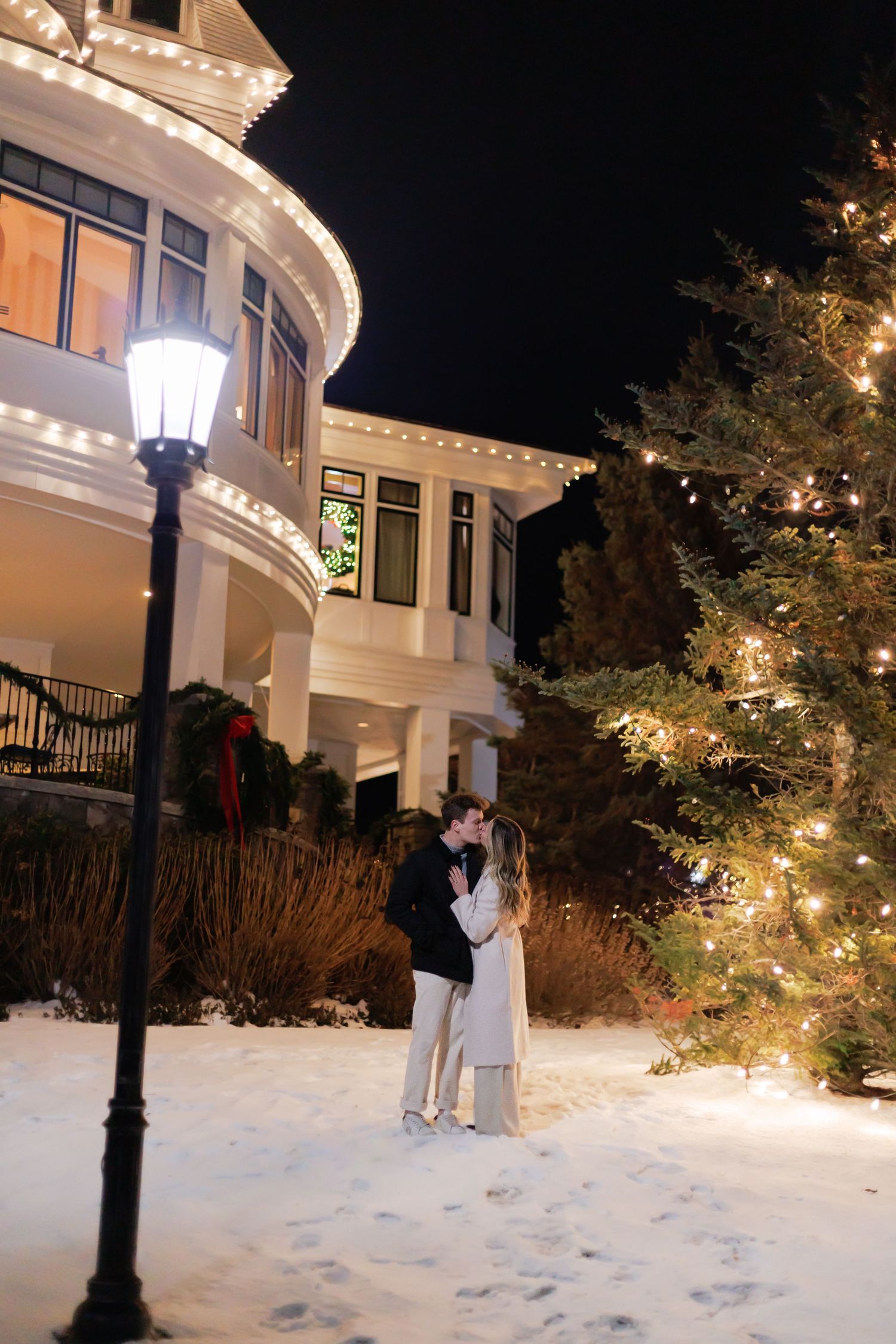Couple embracing in snowy yard at night beside lit Christmas tree and elegant white house with holiday lights.