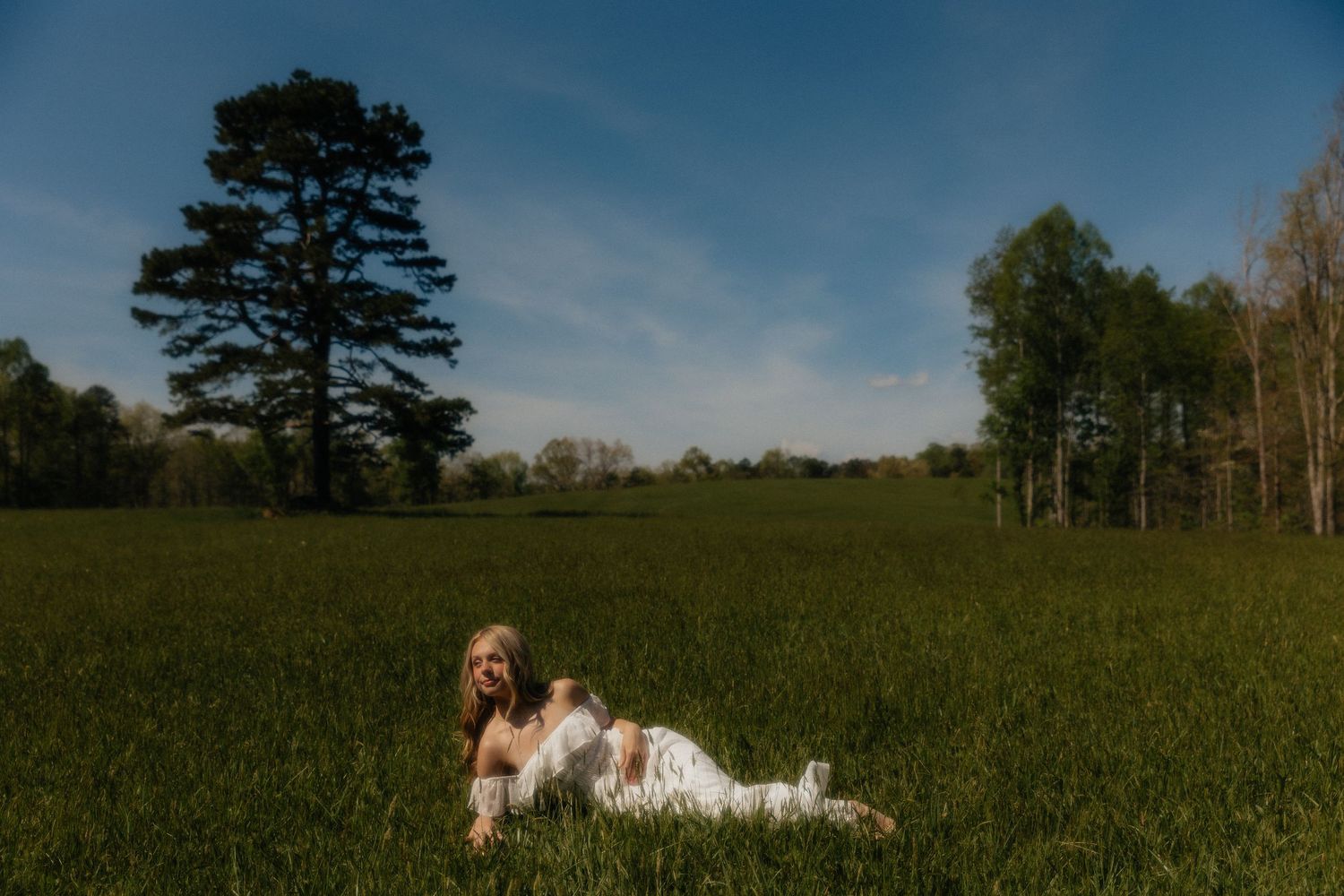 Woman in white dress lying in lush green field with tall trees and blue sky on a sunny day.