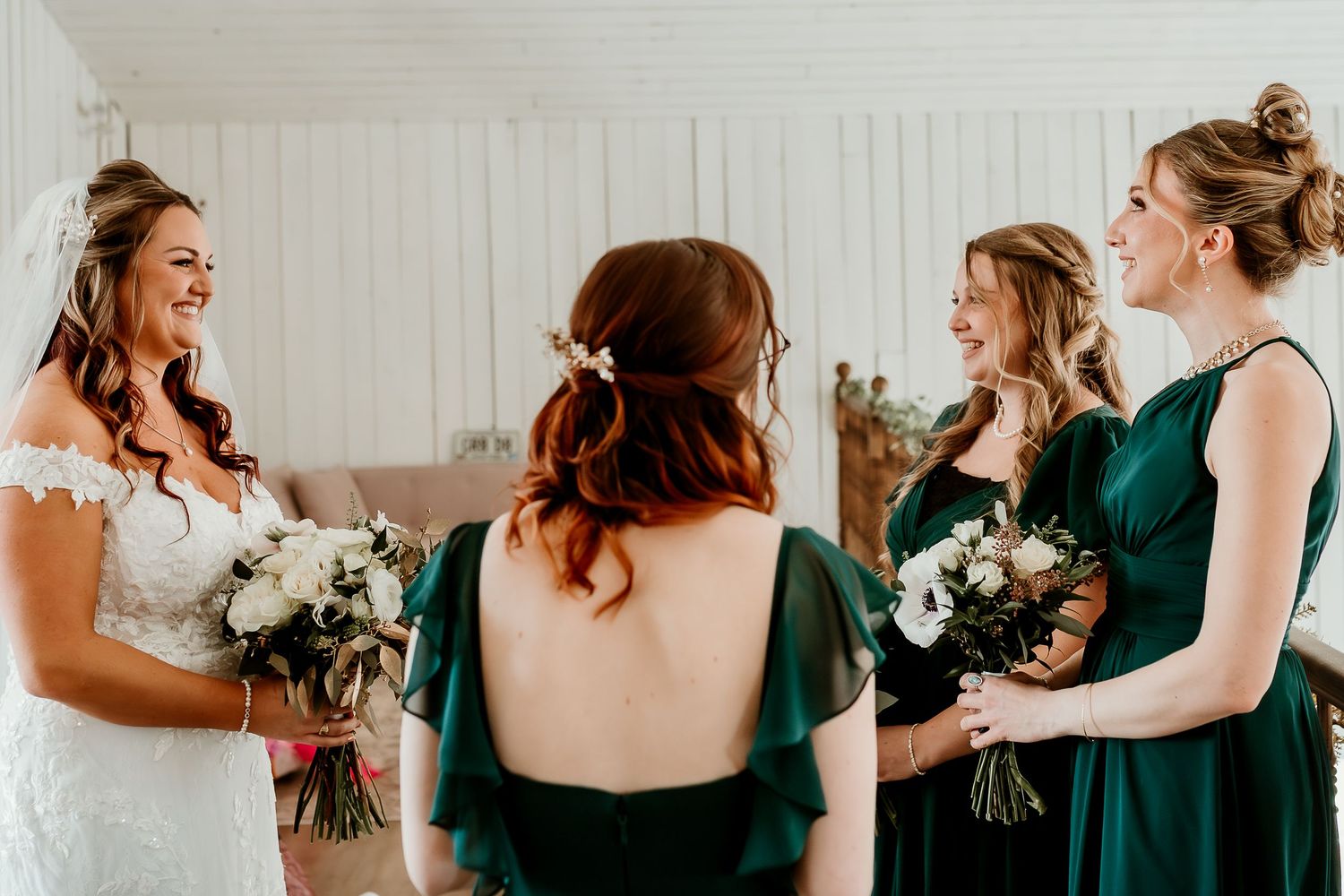Bride in white gown laughs with bridesmaids in emerald green dresses holding white bouquets at wedding.