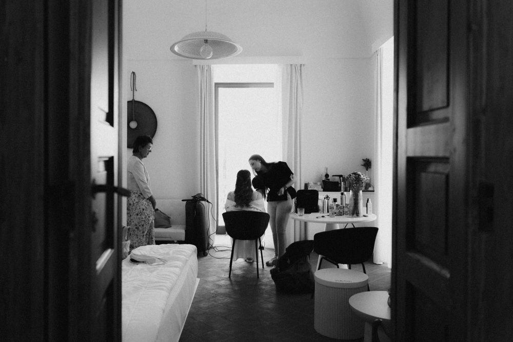 Black and white photo of people in a bright room, with someone getting hair or makeup done near a vanity mirror.